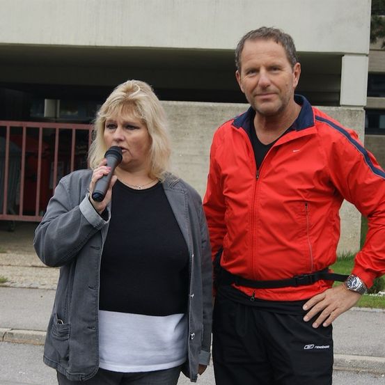 Eine Frau mit blonden Haaren hält ein Mikrofon in der Hand und spricht, während sie neben einem Mann in einem roten Jacken steht. Sie stehen vor einem Gebäude.