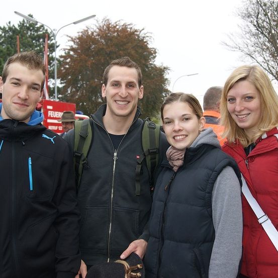 Vier Personen, zwei Männer und zwei Frauen, stehen eng beieinander und lächeln für ein Foto. Sie sind in Winterkleidung gekleidet. Hinter ihnen befinden sich Bäume, ein Feuerwehrauto und eine Straßenlaterne.