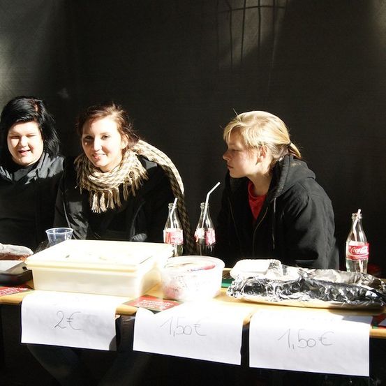 Drei Frauen sitzen an einem Tisch mit Essen und Getränken. Sie lächeln und scheinen sich zu amüsieren. Vor ihnen stehen Schilder mit Preisen.