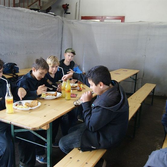 Eine Gruppe von Jungen sitzt an einem Holztisch in einem Raum, isst und trinkt. Ein Junge gießt eine Flasche in einen Becher.