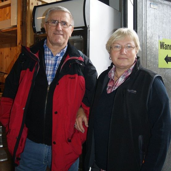 Ein älterer Mann und eine ältere Frau stehen nebeneinander in einem Raum mit Holzwänden und einem Kühlschrank im Hintergrund.