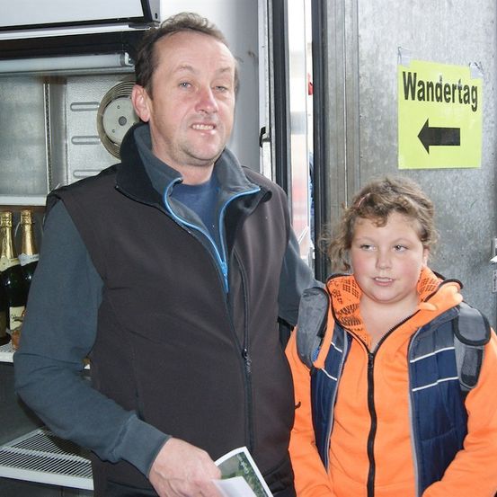 Ein Mann und ein junges Mädchen stehen nebeneinander vor einer Metalltür mit einem gelben Schild, das 'Wandertag' und einen nach links weisenden Pfeil zeigt. Der Mann hält ein Stück Papier. Hinter ihnen ist ein Kühlschrank mit Champagnerflaschen zu sehen.