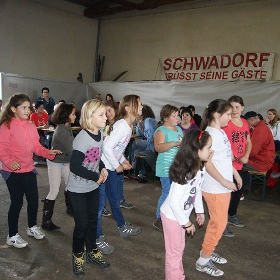 Eine Gruppe junger Mädchen tanzt in einem Raum mit einem Banner, das Schwadorf Grusst Seine Gaste steht.