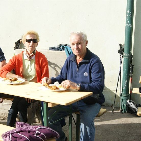 Zwei ältere Menschen sitzen draußen an einem Tisch und essen. Der Mann trägt eine Uhr und die Frau trägt eine Schürze.