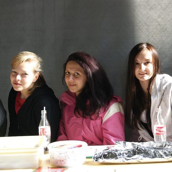 Drei Frauen sitzen an einem Tisch mit Essen und Getränken vor ihnen. Die Frau links hat blonde Haare, die Frau in der Mitte hat dunkle Haare und die Frau rechts hat braune Haare. Es gibt zwei leere Flaschen Coca-Cola auf dem Tisch.