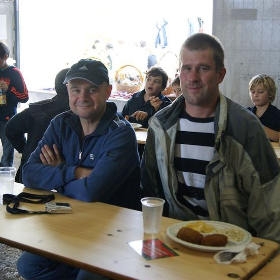 Zwei Männer sitzen an einem Tisch in einer Cafeteria, einer mit verschränkten Armen und der andere lächelt. Es gibt andere Leute, die am Tisch und im Hintergrund sitzen.