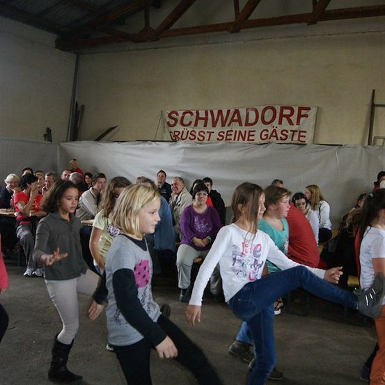 Eine Gruppe von Kindern tanzt in einem Raum mit einem Banner, das 'Schwadorf' über ihnen sagt. Hinter ihnen sitzen Leute auf Stühlen.
