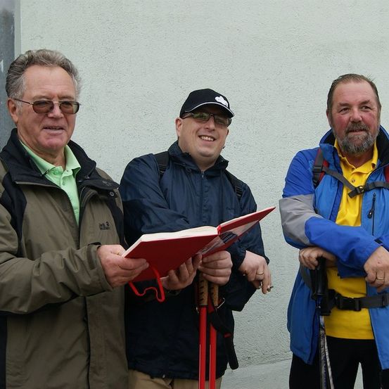 Drei Männer in Outdoor-Kleidung stehen zusammen, einer hält ein rotes Buch, ein anderer lächelt und der dritte hat einen Rucksack.