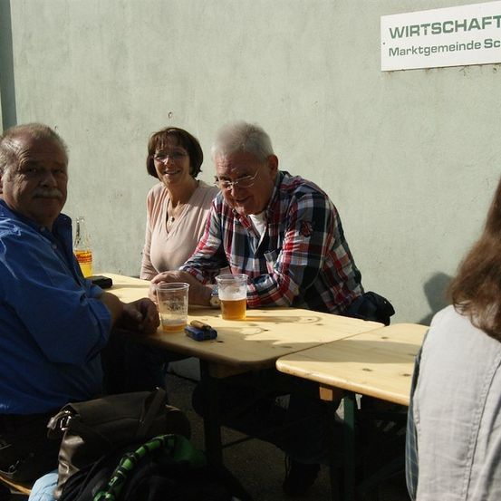 Drei Personen sitzen an einem Tisch, zwei Männer und eine Frau, mit Getränken und einem Schild, das Wirtschaftsmarktgemeinde Schwad besagt, im Hintergrund.