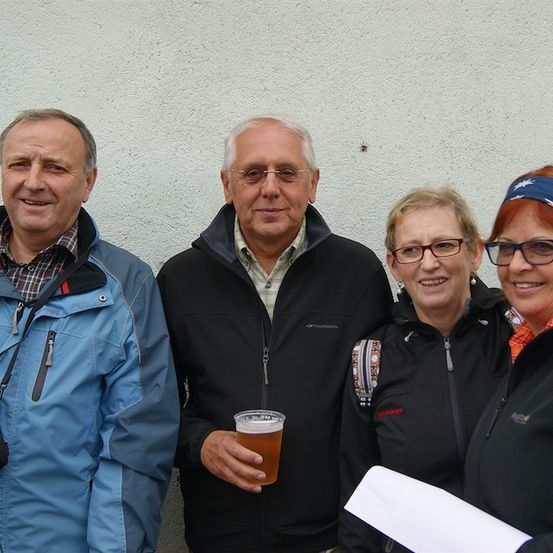 Vier Menschen stehen zusammen und lächeln für ein Foto. Der Mann auf der linken Seite trägt eine blaue Jacke. Der Mann in der Mitte hält ein Glas Bier. Die beiden Frauen tragen Brillen und Jacken.