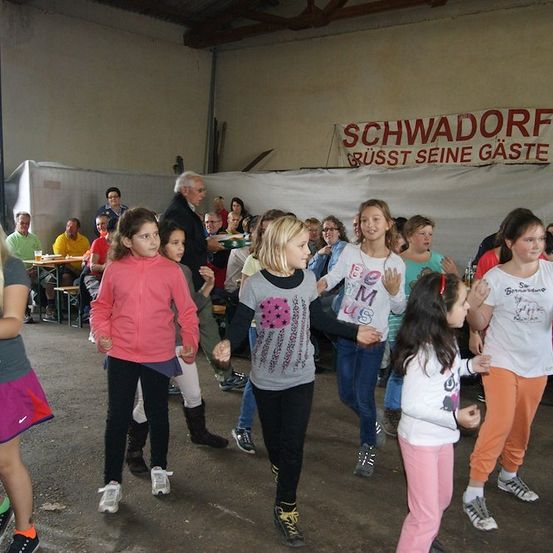 Eine Gruppe junger Mädchen tanzt in einem Raum mit einem Banner, das 'Schwadorf' sagt. Einige Leute sitzen auf Bänken hinter ihnen.
