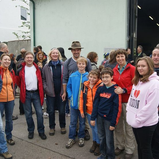 Eine Gruppe von Menschen, einschließlich Erwachsener und Kindern, steht vor einem Gebäude, lächelt und posiert für ein Foto. Sie sind in verschiedenen Jacken und Turnschuhen gekleidet. Das Gebäude hat eine offene Tür und ein Schild.