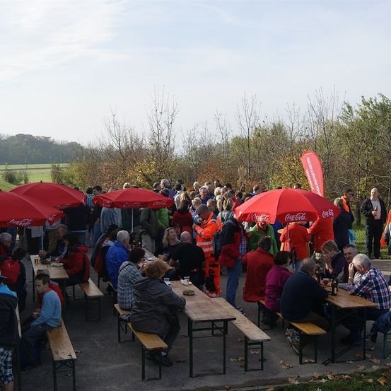 Eine große Gruppe von Menschen ist im Freien für eine Veranstaltung versammelt. Sie sitzen um Tische herum unter roten Sonnenschirmen, einige mit Coca-Cola-Markierung. Die Umgebung ist von Bäumen und einem klaren Himmel umgeben.