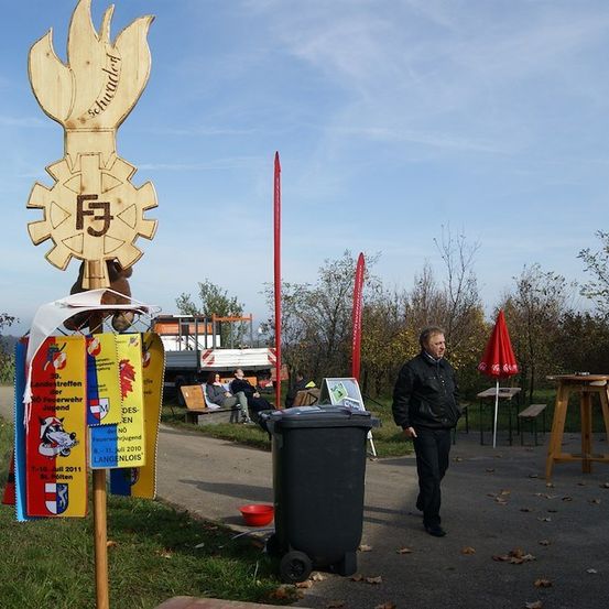 Ein Mann geht an einem Holzschild mit Flammendesign und Text, einem Mülleimer und einem roten Schirm vorbei. Im Hintergrund sitzen Leute auf einer Bank, und ein LKW ist geparkt.