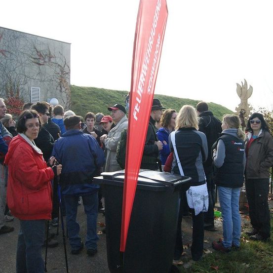 Eine Gruppe von Menschen steht in einer Schlange draußen, einige tragen Jacken und Mützen. Ein roter Banner ist sichtbar, und ein Mülleimer ist in der Nähe. Im Hintergrund zeigt sich ein Gebäude mit Kletterpflanzen und einem grün bewachsenen Hügel.