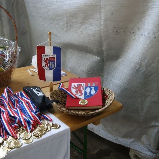 Ein Holztisch zeigt einen Korb mit Medaillen, ein rotes Buch mit Wappen und eine Flagge. Links steht ein Korb mit Gegenständen.