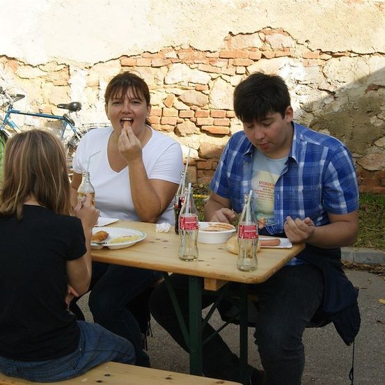 Eine Familie sitzt an einem Picknicktisch und isst. Eine Frau isst einen Hot Dog, ein Mann isst ein Sandwich und ein Mädchen isst einen Salat. Es gibt Flaschen mit Limonade auf dem Tisch.