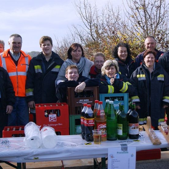 Eine Gruppe von Menschen, einschließlich Feuerwehrleute, steht hinter einem Tisch mit Flaschen und Gläsern. Sie lächeln und posieren für ein Foto.