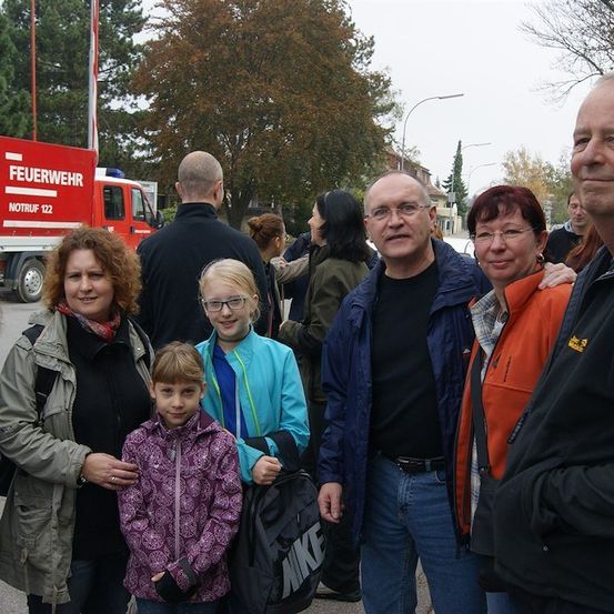 Eine Gruppe von Menschen steht draußen, einige mit Brillen und Jacken, mit einem Feuerwehrauto im Hintergrund.