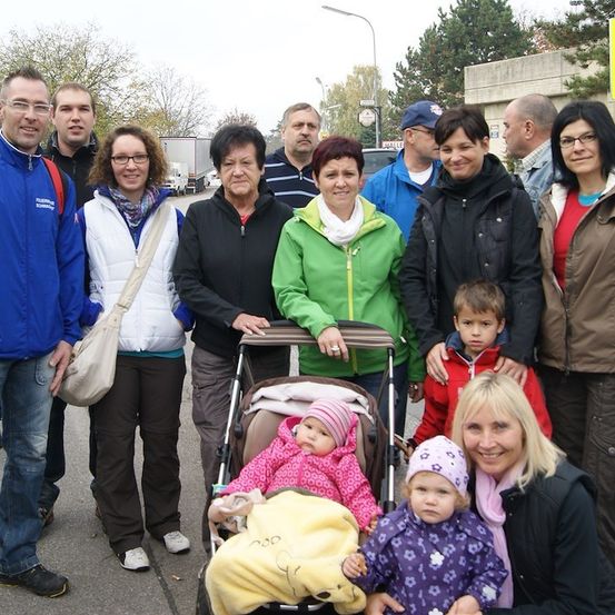 Eine Gruppe von Menschen, einschließlich Erwachsener und Kindern, stehen zusammen auf einem Außengelände, möglicherweise für ein Familienfoto. Einige tragen Winterkleidung.