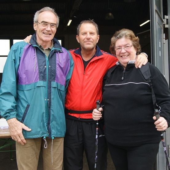 Drei Personen, zwei Männer und eine Frau, stehen lächelnd für ein Foto zusammen. Sie sind in Outdoor-Kleidung gekleidet und halten Trekkingstöcke. Der Mann links trägt eine mehrfarbige Jacke, der Mann in der Mitte eine rote Jacke und die Frau eine schwarze Jacke. Sie stehen vor einem Gebäude mit offener Tür.