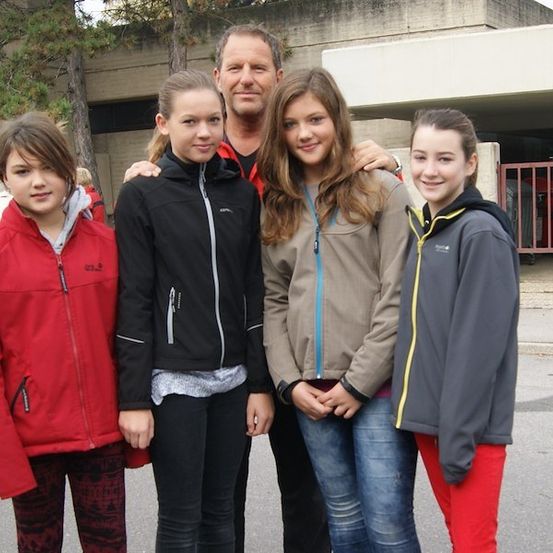Eine Gruppe von fünf Personen, vier jungen Mädchen und einem Mann, stehen zusammen vor einem Gebäude. Sie alle lächeln und posieren für ein Foto. Die Mädchen tragen Jacken in verschiedenen Farben, und der Mann hat seine Arme um zwei der Mädchen gelegt. Hinter ihnen steht ein Gebäude mit einem roten Zaun und einigen Bäumen.