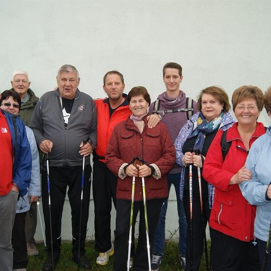 Eine Gruppe von Menschen, die für Outdoor-Aktivitäten gekleidet sind, steht lächelnd vor einer weißen Wand und posiert für ein Foto. Sie halten Skistöcke und einige tragen Brillen.