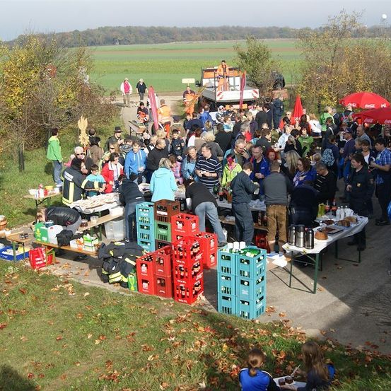 Eine große Menschenmenge versammelt sich im Freien, einige unter Regenschirmen, in der Nähe von Tischen mit Essen und Getränken. Bäume und ein Feld im Hintergrund.