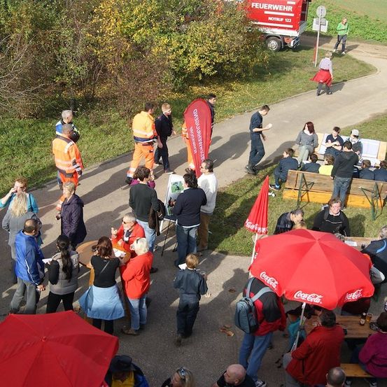 Eine Vogelperspektive einer Menschenansammlung in einem Park, einige stehen und andere sitzen auf Bänken, mit einem Feuerwehrauto und einem Coca-Cola-Regenschirm im Hintergrund.