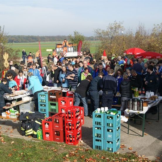 Eine große Gruppe von Menschen versammelt sich um Tische in einer Außenumgebung, möglicherweise für eine Gemeinschaftsveranstaltung. Die Tische sind mit Essen, Getränken und verschiedenen Gegenständen beladen. Einige Personen tragen Jacken und Mützen, während andere Uniformen tragen. Im Hintergrund befindet sich ein Feld und ein Maschinenfahrzeug.