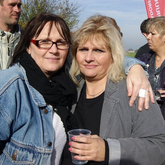 Zwei Frauen stehen eng beieinander, eine hält einen Plastikbecher und lächelt für ein Foto, während eine Gruppe von Menschen im Hintergrund steht.