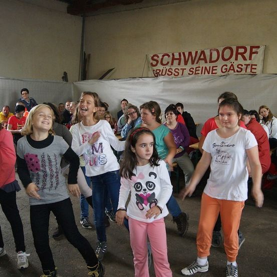 Eine Gruppe junger Mädchen tanzt in einem Raum mit einem Banner, das 'Schwadorf' und 'Grüsst Seine Gäste' steht. Sie tragen Freizeitkleidung und Turnschuhe. Hinter ihnen sitzen Leute auf Stühlen, und einige stehen. Der Raum hat eine weiße Wand und eine Holzdecke.