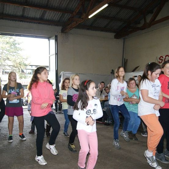 Eine Gruppe junger Mädchen tanzt in einem Raum mit einer großen offenen Tür. Sie tragen Freizeitkleidung und Turnschuhe. Einige Leute stehen hinter ihnen. Es gibt auch Leute, die auf Stühlen im Raum sitzen. Der Raum hat eine weiße Wand und ein Metalldach.