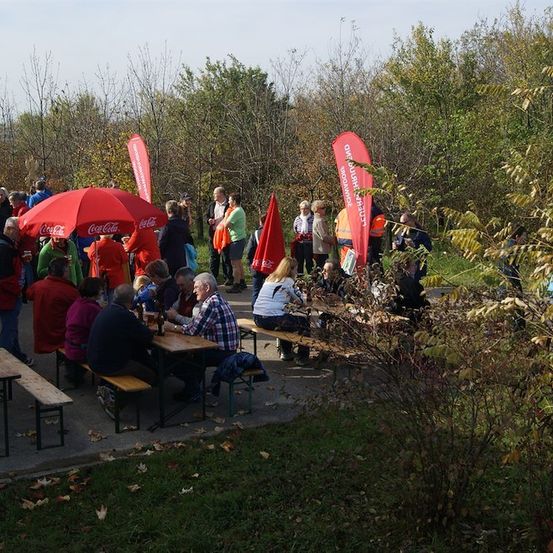 Eine Gruppe von Menschen versammelt sich im Freien um Tische und Bänke, einige unter roten Regenschirmen. Sie sind in verschiedenen Farben gekleidet, und einige tragen orange Westen. Die Umgebung ist von Bäumen und Gras umgeben.
