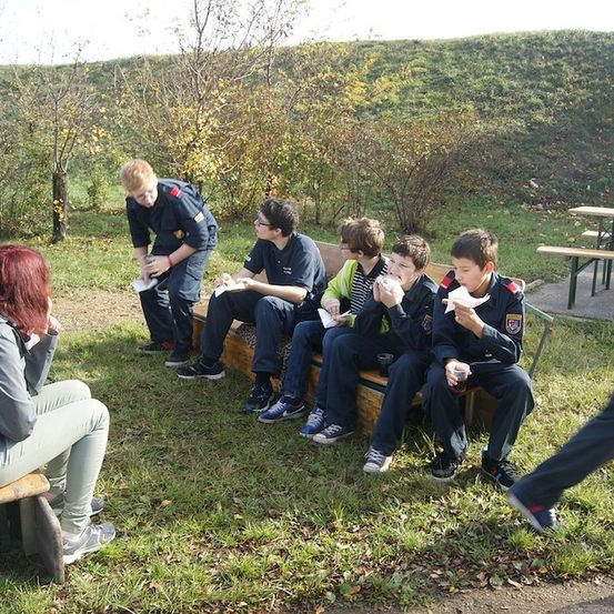Eine Gruppe junger Menschen in Uniformen sitzt auf einer Bank in einem Park, isst Essen und trinkt aus Tassen.