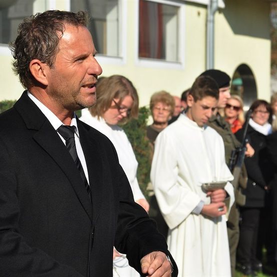 Ein Mann in einem schwarzen Anzug und Krawatte steht vor einer Gruppe von Menschen, möglicherweise bei einer Beerdigung. Hinter ihm hält ein Mann in einem weißen Gewand einen Teller.