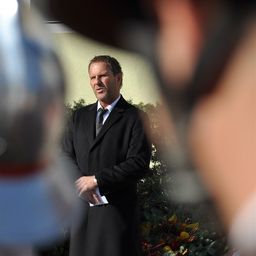 Ein Mann in einem schwarzen Mantel und Krawatte steht im Freien, hält ein Stück Papier in der Hand, mit einem verschwommenen Hintergrund von Pflanzen und einem Kopf einer Person.