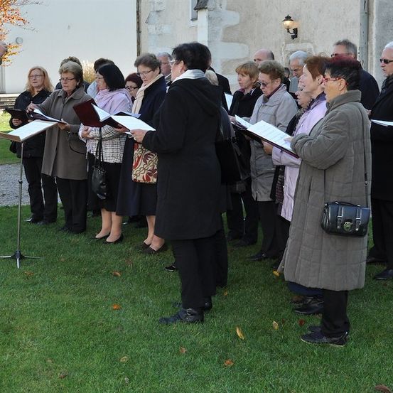 Ein Chor aus Erwachsenen und einem Kind steht auf einem Rasen, hält Notenblätter in der Hand und singt vor einem Gebäude.