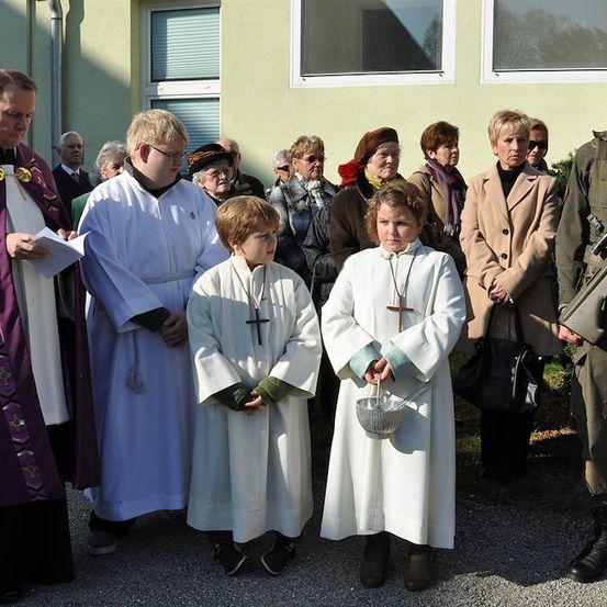 Eine religiöse Zeremonie mit einem Priester und zwei Kindern in weißen Gewändern, einem Soldaten in Uniform und mehreren Erwachsenen vor einem Gebäude.