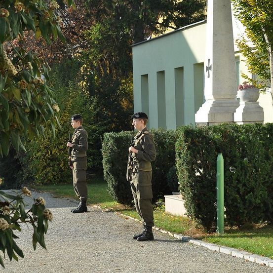 Zwei Soldaten in Uniform stehen vor einem Gebäude mit einem Denkmal dahinter.