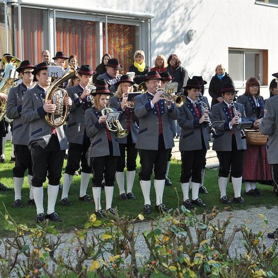 Ein Blasorchester in traditioneller Kleidung spielt auf einem Rasen vor einem Gebäude, mit Zuschauern im Hintergrund.