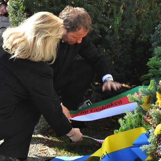 Ein Mann und eine Frau in formeller Kleidung legen eine Schleife auf einen Kranz mit den Worten 'Im Gedenken' vor einem Garten mit immergrünen Pflanzen.