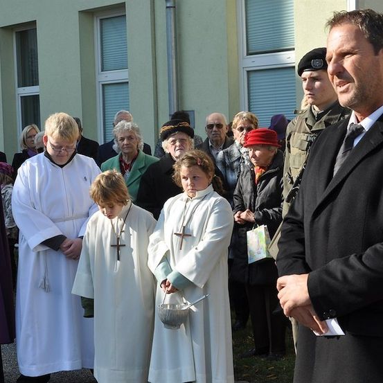 Eine Gruppe von Menschen, einschließlich eines Mannes in einem schwarzen Anzug, versammelt sich vor einem Gebäude. Zwei Kinder in weißen Roben halten eine silberne Schüssel.