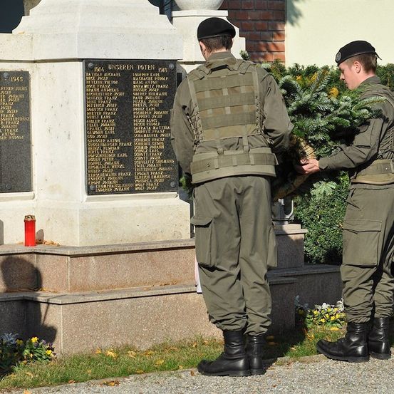Zwei Soldaten in grünen Uniformen stehen vor einer Gedenktafel und halten einen Kranz. Die Tafel hat Namen und Daten, und eine rote Kerze ist in der Nähe.