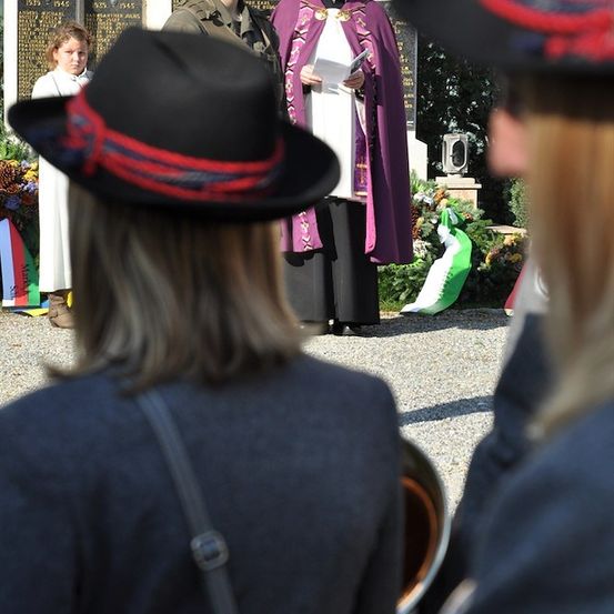 Eine feierliche Zeremonie mit einem Priester und Militärangehörigen vor einem Denkmal mit Tafeln und Blumen.