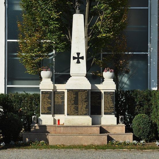Ein weißes Denkmal mit einem Kreuz und Tafeln steht vor einem Backsteingebäude mit Glasfenstern. Zwei kleine Bäume mit rosa Blumen stehen auf beiden Seiten des Denkmals.