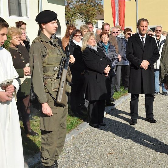 Eine Gruppe von Menschen, darunter ein Soldat und ein Mann in einem schwarzen Anzug, stehen in einer Reihe auf einem Kiesweg. Ein Junge in einem weißen Gewand hält einen Becher.