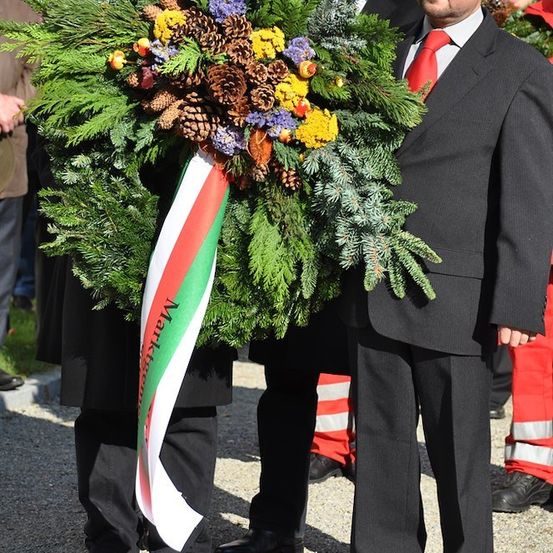 Ein Mann in einem schwarzen Anzug, der einen Kranz mit einem ungarischen Flaggenband hält, steht neben einem anderen Mann in einem Anzug und einer Person in rot-weißer Uniform.