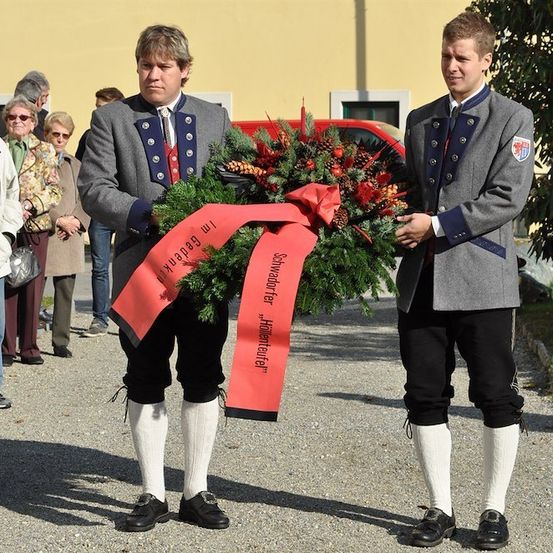 Zwei Männer in traditionellen Uniformen halten einen Kranz mit einem roten Band und stehen vor einem Gebäude mit Zuschauern im Hintergrund.