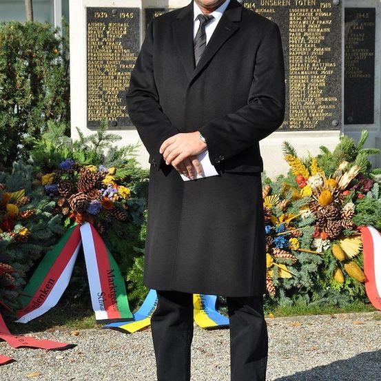 Ein Mann steht vor einer Gedenkwand mit Namen und Blumen dahinter. Er ist in einem schwarzen Anzug gekleidet und hält ein Stück Papier in der Hand.